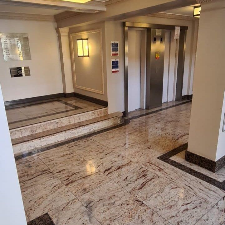Our team of professional cleaners wearing uniforms and gloves, cleaning a communal area with modern equipment, including a vacuum cleaner, mop, and bucket. The area is clean and well-maintained, with floors and windows shining, and trash cans neatly placed in the corner.